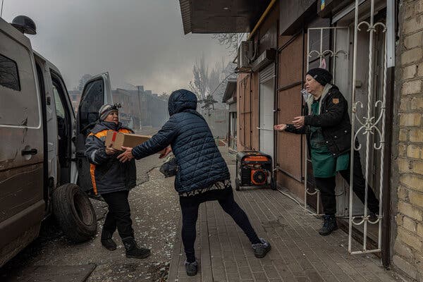 Žena vedľa dodávky podáva krabicu inej osobe vedľa domu s otvorenou bránou.