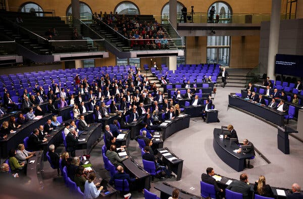 Ľudia sediaci na fialových stoličkách v zákonodarnej sále a obklopujúci dvoch ľudí sediacich za stolom.